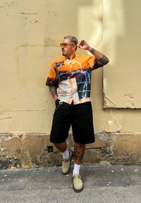 Μακρυμάνικη, πορτοκαλί και μαύρη πουκαμίσα με μοτίβο από συρματόσχοινο, συνδυασμένη με μαύρο σορτς και μπεζ παντόφλες.