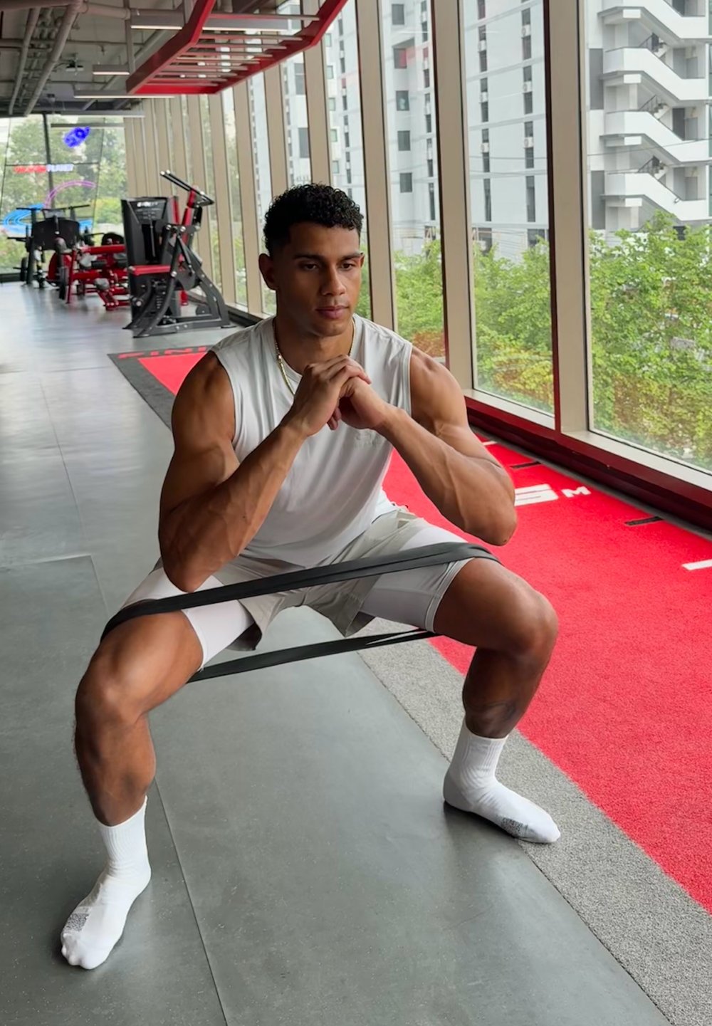 Bande de résistance utilisée lors d'un exercice de squat. Individu portant un débardeur blanc et un short clair sur un sol de salle de sport avec des accents rouges.