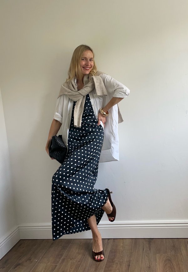Kvinde smiler og poser indendørs iført en sort og hvid polkaprikket kjole, hvid skjorte, beige sweater, holder en sort clutch.