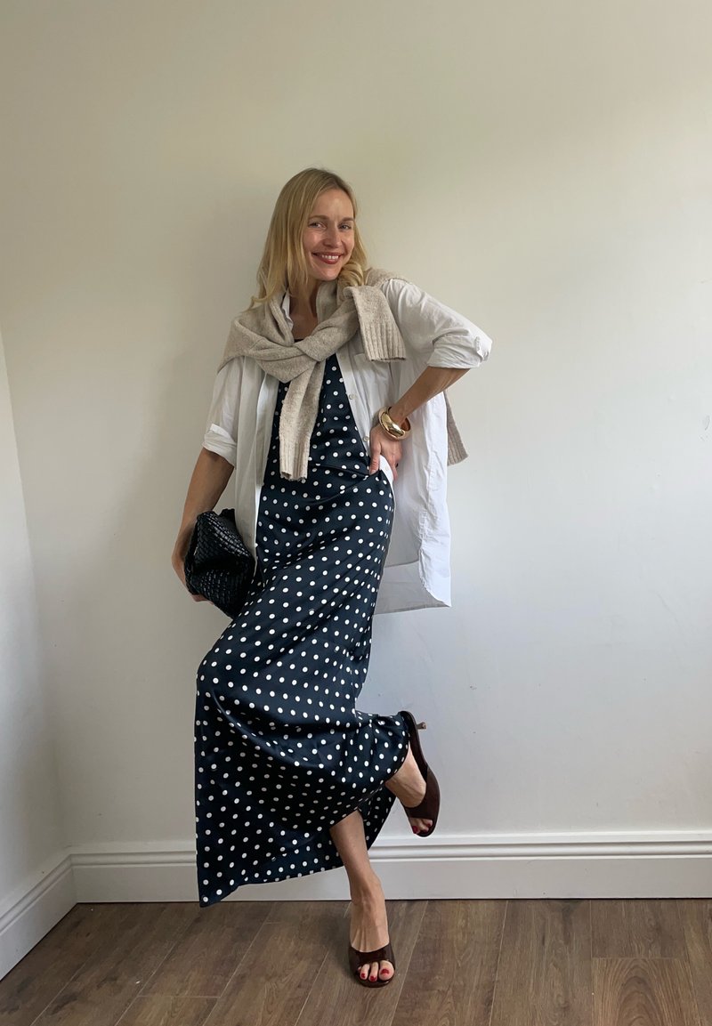 Femme souriante posant à l'intérieur, portant une robe à pois noir et blanc, une chemise blanche, un pull beige, tenant une pochette noire.