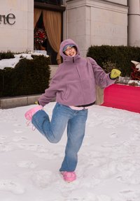 Personne en veste à capuche violette, jean bleu et chaussures roses posant de manière ludique sur un sol enneigé près d'un bâtiment avec des arbustes et une couronne festive.