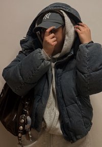 Geacă groasă bleumarin cu fermoar și glugă; hanorac gri dedesubt; șapcă gri; geantă maro cu accesorii metalice vizibile și breloc.