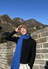 Mlada oseba, oblečena v črn pulover z zadrgo in modro šal, z rjavo pleteno kapo, stoji pred Velikim zidom in gorami.