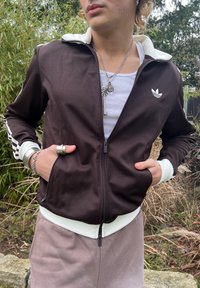 Veste de sport marron avec col et poignets blancs, fermeture éclair, poches latérales, et accents rayés noirs et blancs sur les manches.