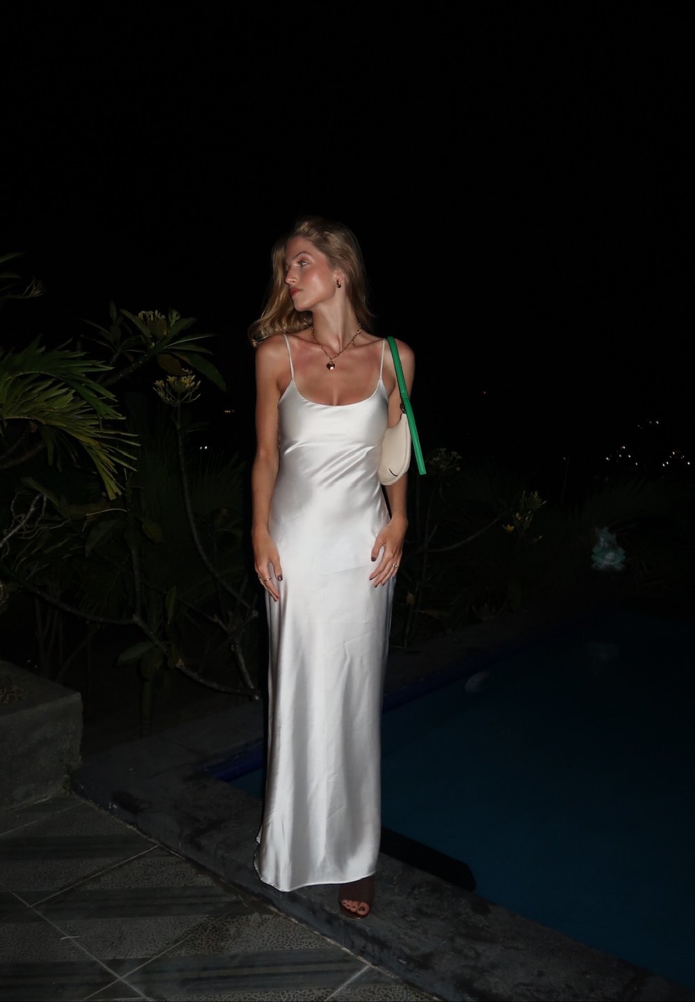 Seidenweisses, ärmelloses, bodenlanges Kleid in hellem Silber; hat eine taillierte Silhouette und einen subtilen Glanz. Ergänzt durch eine hellfarbige Handtasche.
