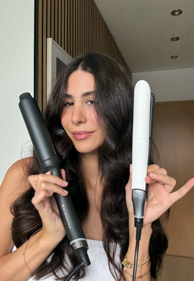 Se sostienen dos herramientas de peinado: un rizador negro con un acabado suave y una plancha blanca con un diseño elegante, ambas con empuñaduras texturizadas.
