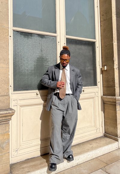 Traje gris oversized con pantalones de pierna ancha, camisa blanca y corbata marrón. De pie contra una pared de color claro, usando gafas de sol y zapatos negros.