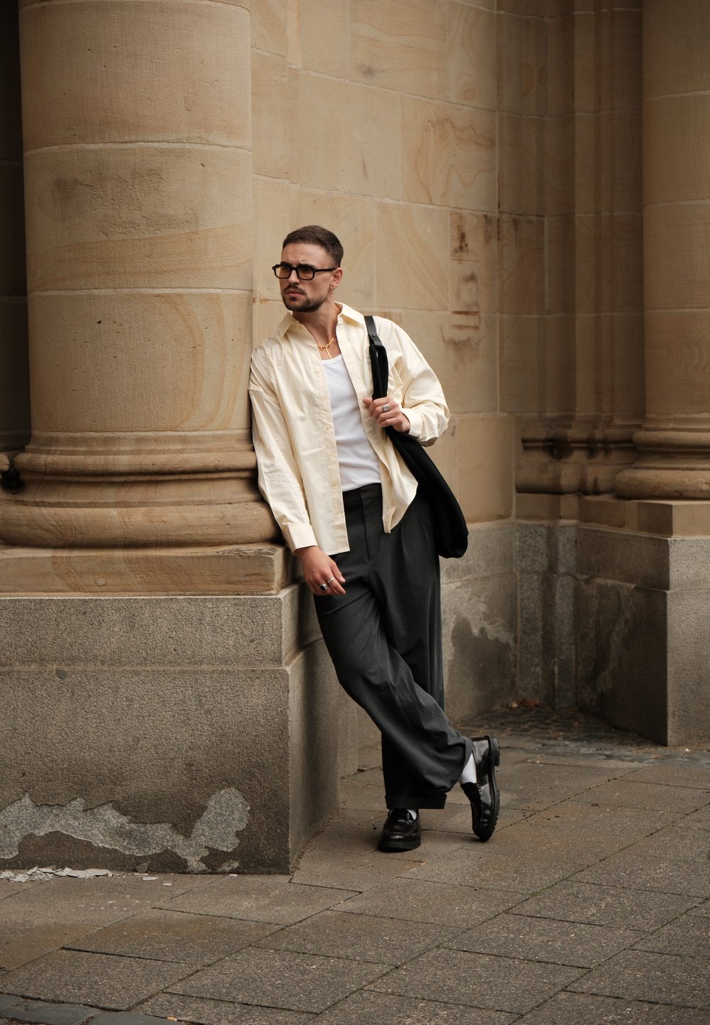 Hellbeige gestreiftes Hemd, weißes Tanktop, graue weite Hose, schwarze Schuhe, ergänzt mit Ringen und einer Brille, angelehnt an eine Steinsäule.