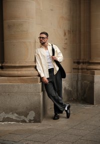 Hellbeige gestreiftes Hemd, weißes Tanktop, graue weite Hose, schwarze Schuhe, ergänzt mit Ringen und einer Brille, angelehnt an eine Steinsäule.