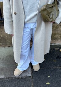 Personne portant un manteau beige, un pantalon blanc et des chaussures crème, tenant un sac à main vert olive texturé, debout sur un trottoir pavé.