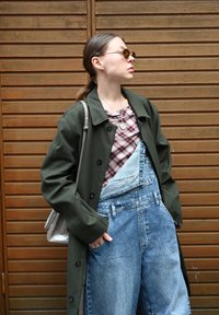 Olijfgroen lange jas, geblokt topje, blauwe denim tuinbroek. Accessoires zijn een zilveren ketting en ronde zonnebril. Glanzende metallieke tas.