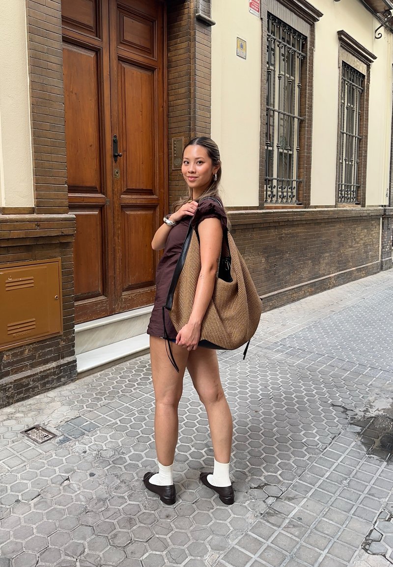 Femme en robe marron portant un grand sac tissé couleur tabac, debout sur un trottoir à motifs devant un bâtiment en brique et beige avec des portes en bois.