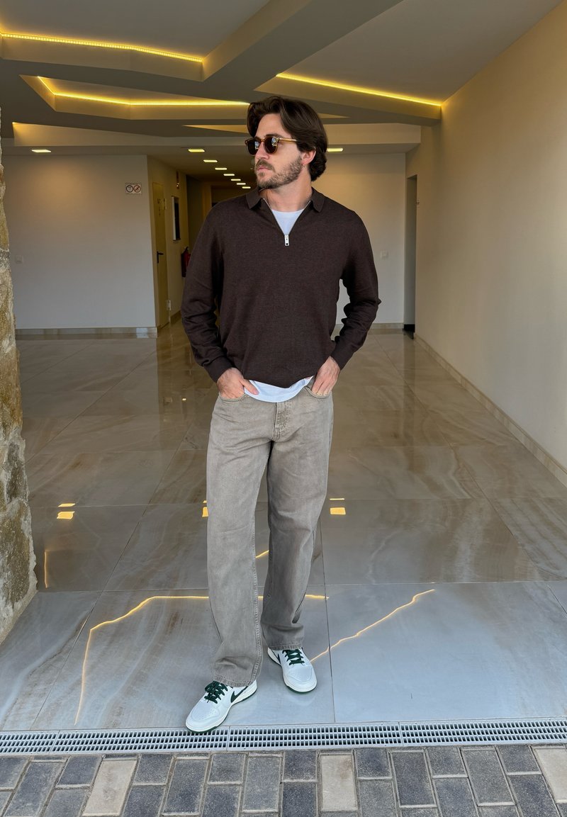 Bruine ritstrui over een witte shirt, beige broek en witte sneakers met groene accenten. De setting heeft een gladde, reflecterende vloer.