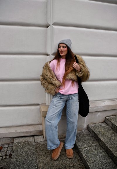 Sudadera rosa, abrigo de piel sintética, jeans de pierna ancha azul claro, botas beige, bolso de punto negro, gorro gris, de pie contra una pared blanca texturizada.