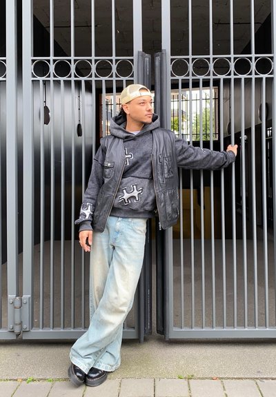 Chaleco de cuero negro sobre una sudadera gris con diseño de piezas de rompecabezas, jeans holgados azul claros y zapatos negros. Puerta de metal al fondo.
