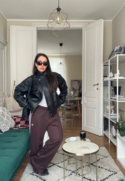 Mujer con chaqueta de cuero negra, gafas de sol y pantalones Adidas marrón oscuro, de pie con las manos en las caderas en una sala de estar moderna.