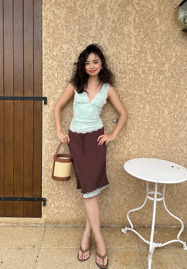 Jonge vrouw met krullend haar in een lichtgroene kanten top en een bruine rok houdt een gevlochten tas vast, staande bij een beige gestructureerde muur en een witte metalen tafel.
