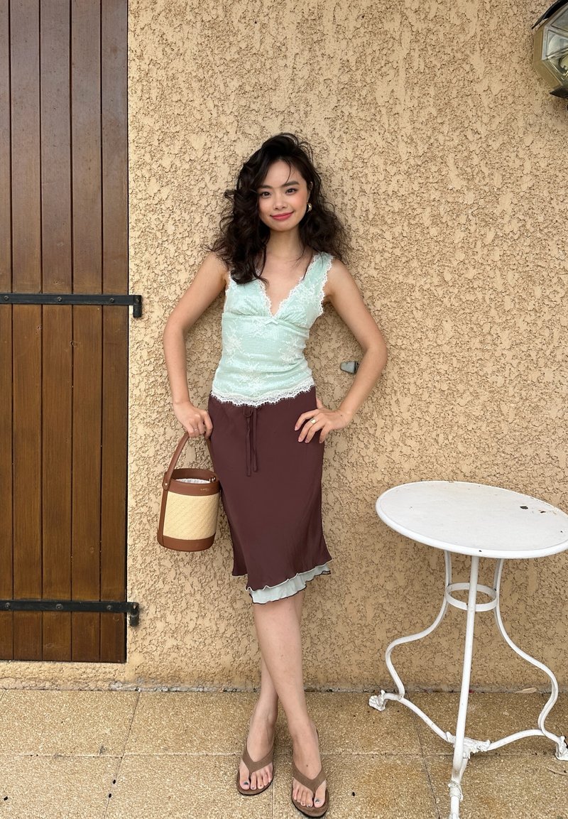 Young woman with curly hair in pale green lace top and brown skirt holds a woven bag, standing by a beige textured wall and white metal table.
