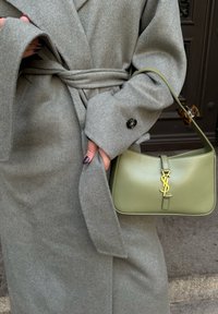 Grau wollener Mantel mit gebundenem Taillengurt, ausgestattet mit großen Knöpfen. Olivgrüne Leder-Schultertasche mit goldenem YSL-Logo auf der Vorderseite.