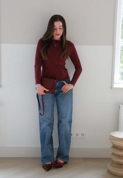 Top de manga larga en color burdeos combinado con jeans acampanados de un azul claro. Detalles notables incluyen un bolso de hombro marrón y tacones rojos de punta. Fondo liso.