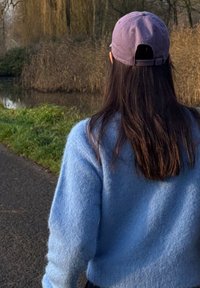 Pull bleu clair en tissu duveteux et casquette de baseball lavande. La scène montre un sentier le long d'un plan d'eau, entouré de hautes herbes.