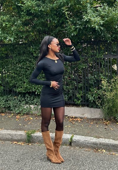Vestido negro de manga larga con fruncidos y abertura lateral, combinado con botas de ante beige hasta la rodilla. Los accesorios brillantes incluyen un collar de cadena y pulseras.