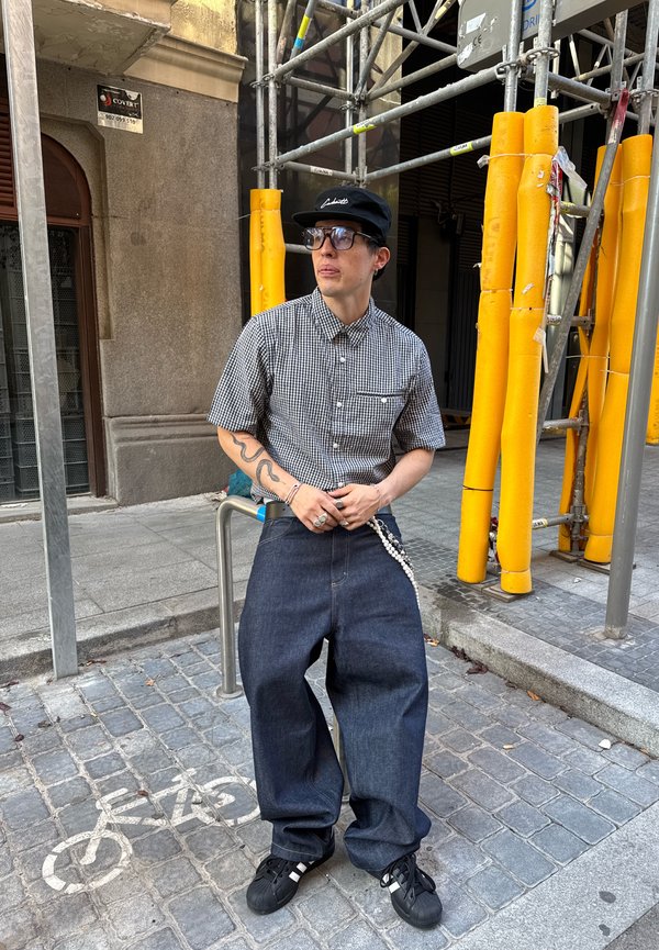 Camisa de botões, de mangas curtas, xadrez, com um bolso, combinada com jeans escuros largos. Usada com sapatilhas pretas e um boné preto.