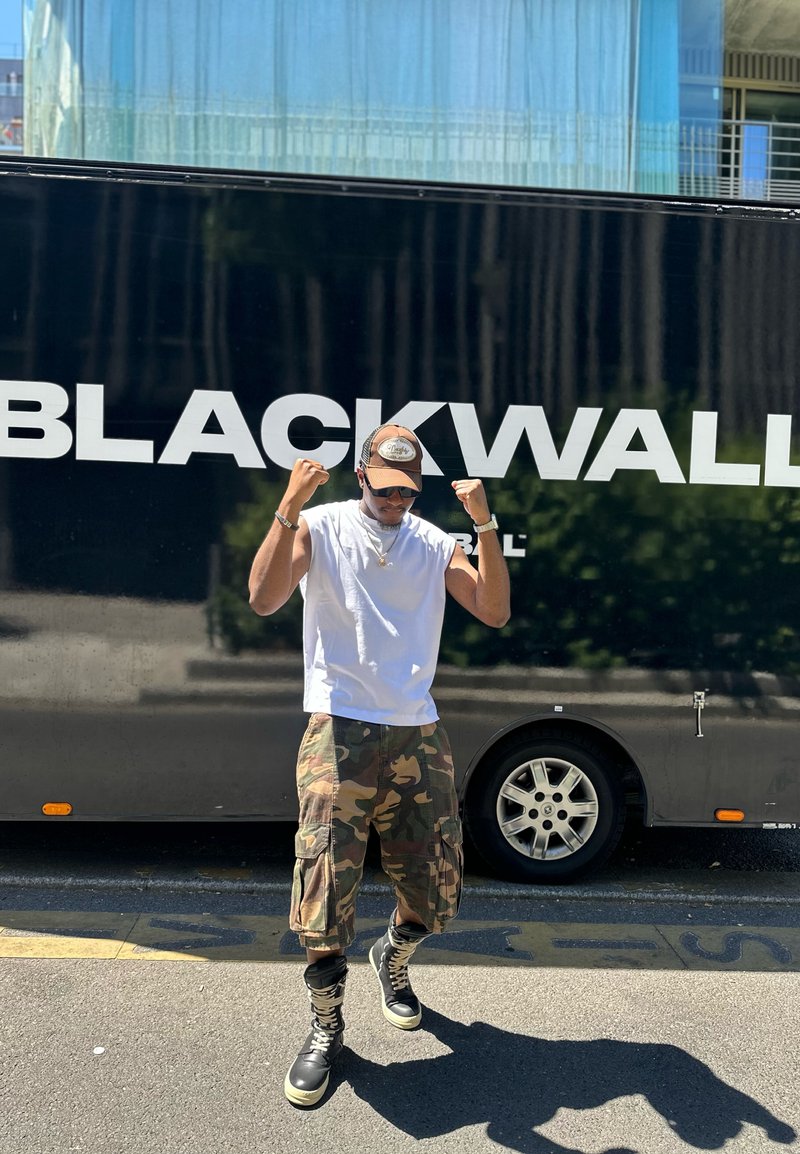 Homme portant une casquette, des lunettes de soleil, un débardeur blanc et un pantalon camouflage pose avec les poings levés devant un véhicule noir avec le texte "BLACKWALL".