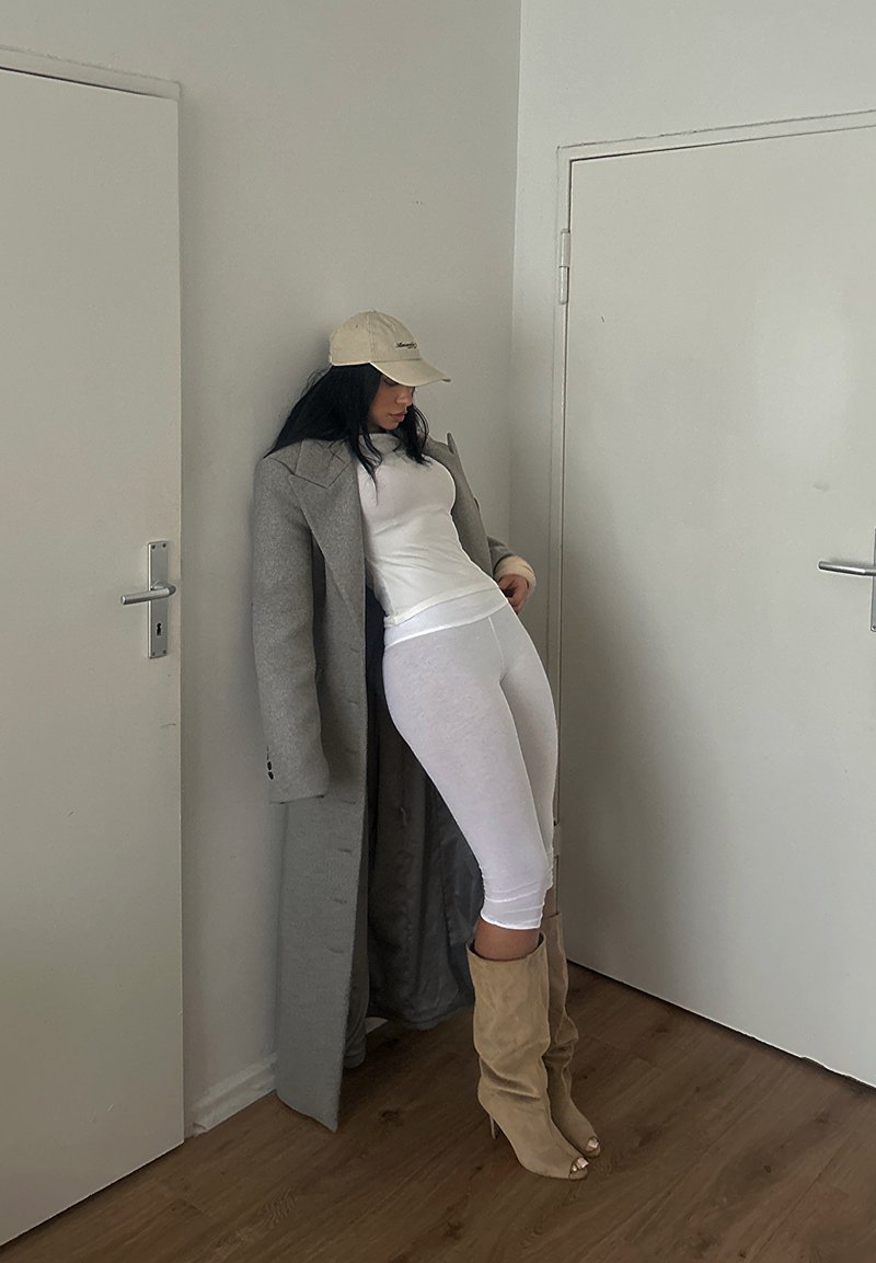 Femme portant un long manteau gris, un haut blanc, des leggings blancs, des bottes beige et une casquette beige, appuyée contre un mur blanc entre deux portes.