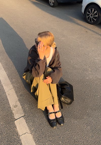 Persona agachada en la acera, vistiendo una sudadera oscura y una camisa clara, pantalones verde olivo y zapatos negros con correas, sosteniendo una bolsa.