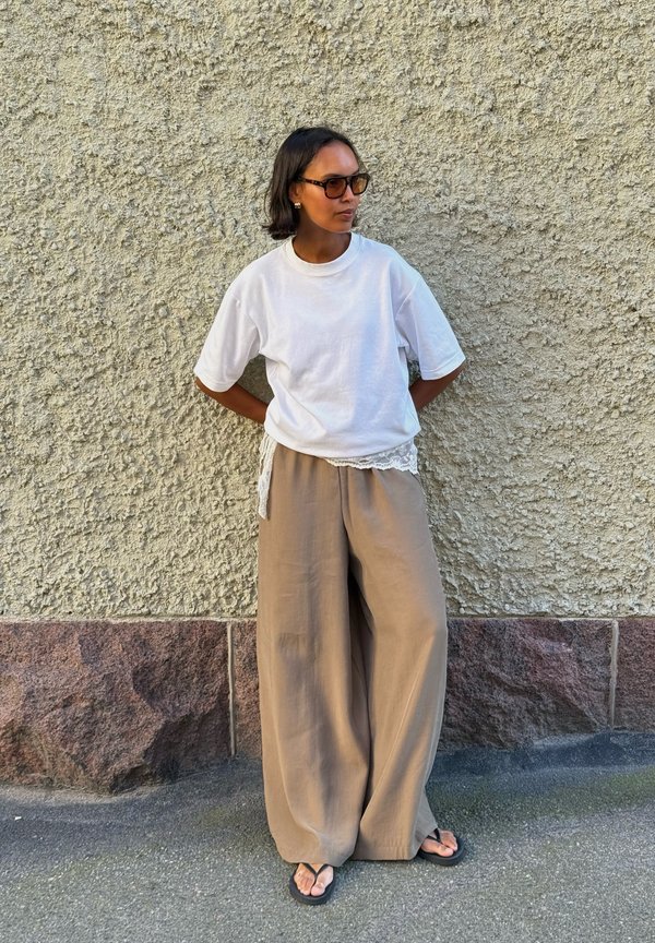 Persona con camiseta blanca extragrande, pantalones beige de pierna ancha, chanclas negras y gafas de sol, de pie contra una pared texturizada con las manos detrás de la espalda.