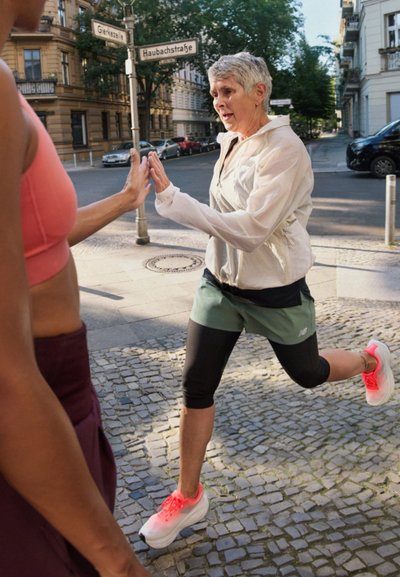Dos mujeres en ropa deportiva, una con un sujetador deportivo rosa y la otra con una chaqueta ligera y pantalones cortos verdes, se chocan las manos en una calle de la ciudad.