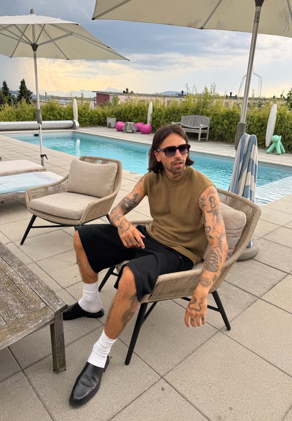Homme avec des tatouages portant des lunettes de soleil, un pull sans manches, un short et des chaussures noires, assis au bord d'une piscine sous des parasols par une journée nuageuse.