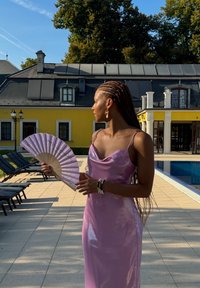 Abito di raso rosa con scollatura a barchetta, che tiene un ventaglio lilla. Capelli intrecciati, orecchini a cerchio e braccialetti a strati. Patio con piscina sullo sfondo.