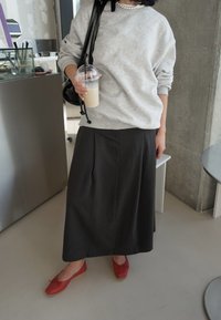 Siva prevelika bunda, nabran temno krilo in rdeče balerinke. Drži ledeno pijačo v prozornem kozarcu. Črna torba za ramo z detajli.