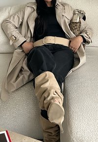 Beżowy trencz na czarnym topie, czarnych dżinsach i beżowych botkach. Beżowy pasek podkreśla talię. Torebka w wzór spoczywa na ramieniu.