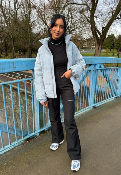 Chaqueta de pana azul clara con un cuello alto, combinada con pantalones negros que llevan un logo. Zapatillas blancas y azules completan el conjunto.