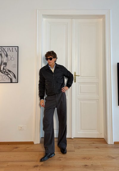 Chaqueta bomber negra con bolsillos frontales, camisa azul claro, pantalones anchos oscuros y gafas de sol, posando cerca de una puerta blanca y una obra de arte enmarcada.