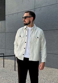 Gaišas denima jakas ar divām pogām priekšējās kabatās, melnas pogas, valkāts virs baltas T-kreklu, saskaņots ar tumšām biksēm.