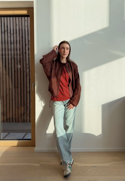 Joven mujer con chaqueta marrón, camiseta de rayas rojas y jeans claros apoyada contra una pared blanca iluminada por el sol cerca de una puerta de madera en el interior.
