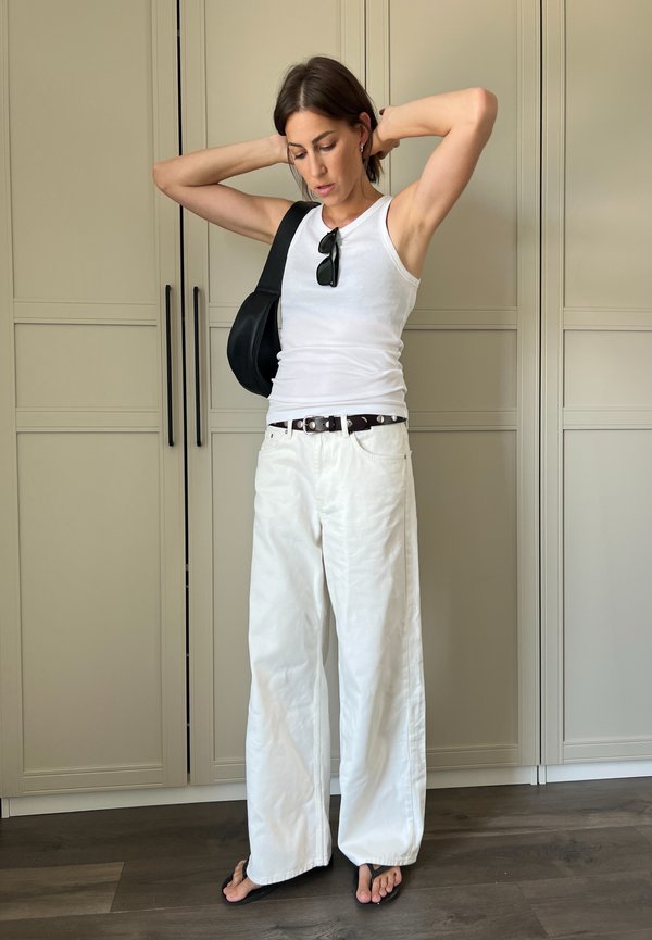 Pantalones de lino blanco de pierna ancha, top ajustado blanco, bolso negro y chanclas. Texturas suaves con un diseño casual. Suelo de madera y armarios en tonos neutros.