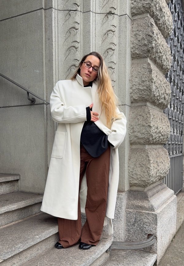 Femme aux longs cheveux blonds et lunettes, vêtue d'un manteau blanc, d'une chemise noire et d'un pantalon marron, debout sur des marches en pierre près d'une façade de bâtiment ornée.