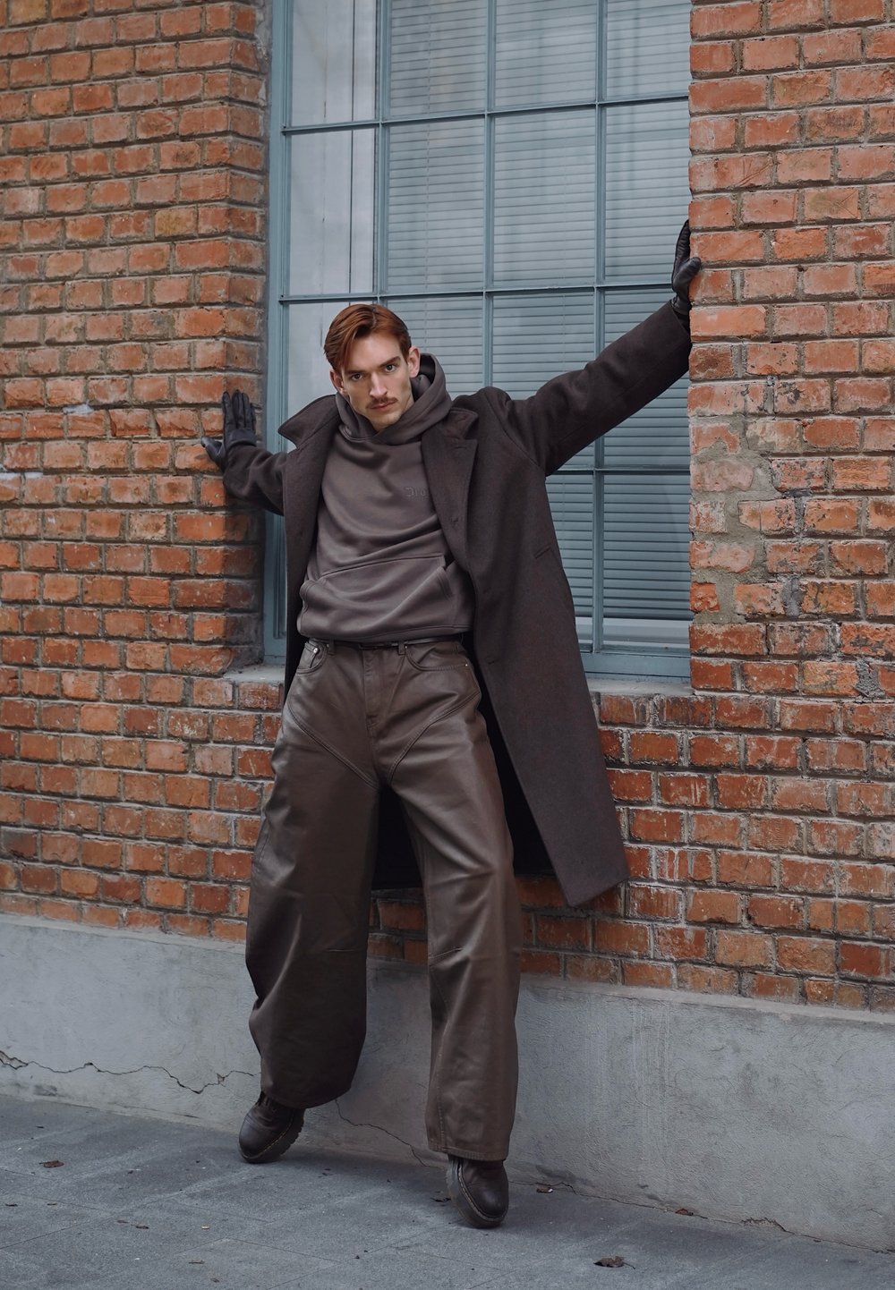 Homme portant un pantalon en cuir foncé, un long manteau, des gants et un sweat à capuche, posant avec les bras étirés contre un mur de briques sous une grande fenêtre.