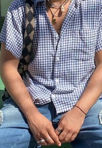 Camisa de manga curta com botões, na cor branca, com padrão de xadrez azul e preto. Usada com jeans desgastados azuis, apresentando vários anéis e colares.