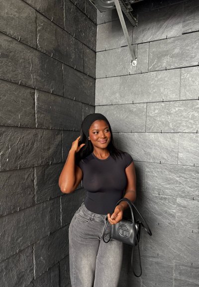 Top ajustado negro de manga corta, jeans grises de cintura alta y un pequeño bolso negro con logo. Fondo de pared de piedra texturizada de color gris oscuro.