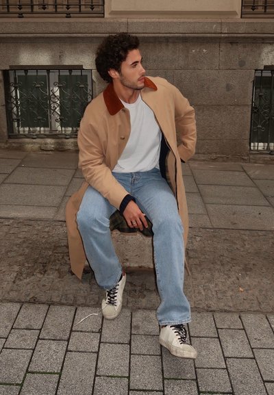 Abrigo beige con collar marrón, camiseta blanca, vaqueros azules y zapatillas blancas. Sentado en un banco de piedra, con un suelo texturizado y un fondo estampado.