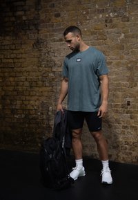 Γκρι-πράσινη μπλούζα με μικρό λογότυπο, μαύρη αθλητική βερμούδα, λευκά παπούτσια και μαύρο σακίδιο σε σκοτεινό πάτωμα μπροστά σε τοίχο από τούβλα.