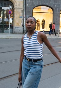 Jauna sieviete svītrainā bezpiedurkņu krekla un džinsos, ejot pa pilsētas ielu ar tramvaja sliedēm, nēsā somu, fona attēlā redzami veikali un gājēji.