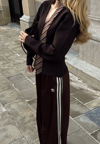 Brūns īss žakete ar garām piedurknēm un rakstaina kaklasaiti. Tumši brūnas bikses ar baltām svītrām un Adidas logotipu uz kājas.