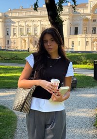 Žena drži prugastu šalicu kave, knjigu i tkanu torbu. Na sebi ima smeđu majicu bez rukava preko bijele košulje i sive hlače.